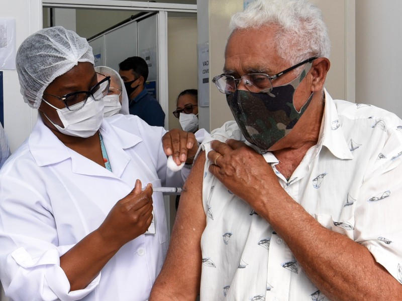 Foto de vacinação contra a Covid-19 num posto de saúde. Uma profissional da saúde negra, com máscara, jaleco e touca brancos aplica a vacina no braço de um homem negro, que está segurando a manga da camisa para a aplicação da seringa. Ele usa óculos e máscara com estampa de camuflagem